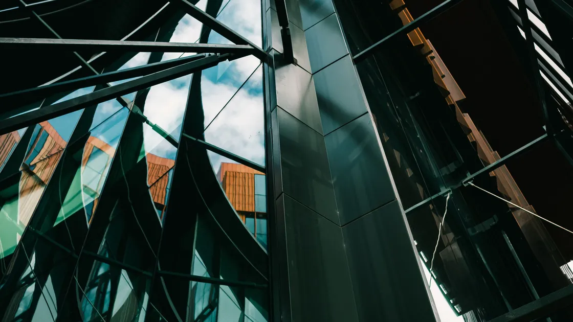 A glass and dark metal facade seen from below, with sky and neighbouring buildings reflected in the angled panes